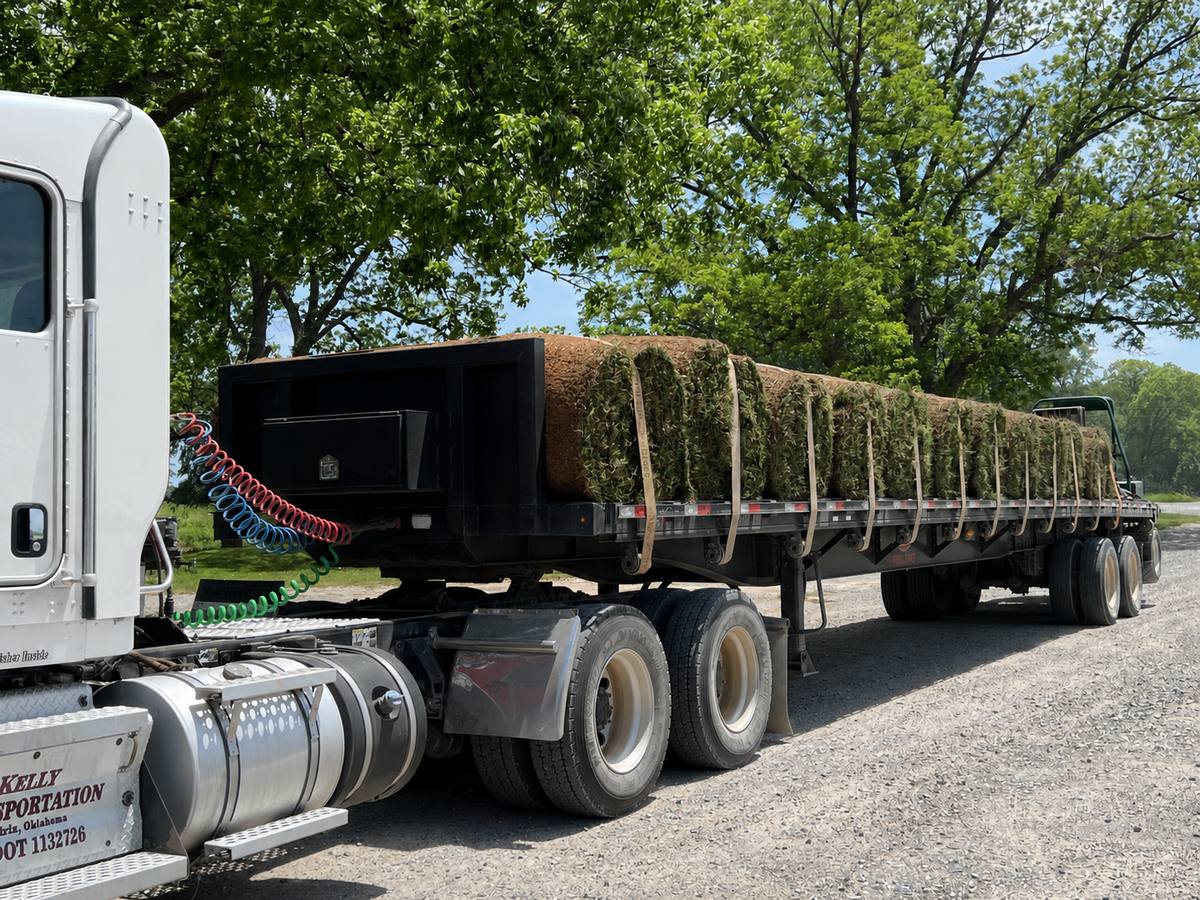 Kelly Transportation flatbed loaded with netted big rolls under the trees